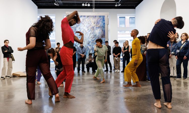 Six dancers are captured mid-motion in an open gallery space, a large blue abstract painting hanging behind them in the distance, and with about twenty people forming a half-circle around them. The dancers wear various costumes of a solid color--orange, black, red, gray--and all face different directions, moving in individual ways. One hugs themself, one twists their upper body to the side; one arches their chest upwards, another stands in profile with bent knees. The audience of all ages stands around them attentitively, some holding programs, cameras, or phones.