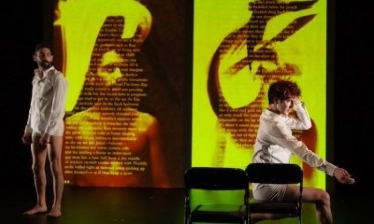 Two male dancers wear white boxer briefs and white button up shirts. One is standing off to the left, arms at his sides looking over to the other dancer, on the right, seated in one of two black folding chairs that face upstage. His left leg is behind the chair, his right leg out in front, foot flat on the floor. He reaches his right hand out, palm turned down, and his left brushes past his face into his hair. Behind them the left side of a split screen has the image of a shirtless man, with a wide bright streak of yellow light on the upper edge. Over the image are lines of text, unreadable from this distance. On the right screen another man is obscured by the seated dancer. The image is mostly bright yellow with black brush strokes, and is also covered in unreadable lines of text.
