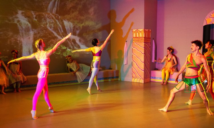 Eight dancers pose within a brightly-lit room, each wearing colorful costumes and with painted pillars around them evoking ancient Greece. Four of them sit or recline along the wall upstage, wearing flowers in their hair and adorned in tulle skirts. In front of them are two women in profile, balanced on the balls of their feet and extending their right arms as they look up. Facing them and to the right, two more dancers stand with their bent thighs wide apart, their curved arms away from their sides like warriors. On the wall upstage of all of them is a tranquil video image of two waterfalls.