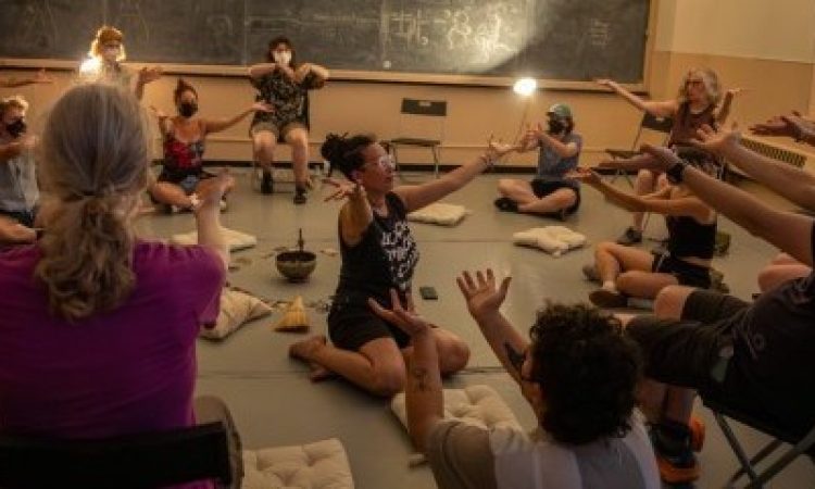 Severin Blake sits on the floor in the middle of a studio space with their arms outstretched toward an audience member. Blake is surrounded by a group of people sitting on chairs and pillows with their arms outstretched as well. There is a singing bowl, some cards, and a broom head sitting on the floor at the center of the space. The studio is dimly lit with two clip lights that are mounted on the wall.