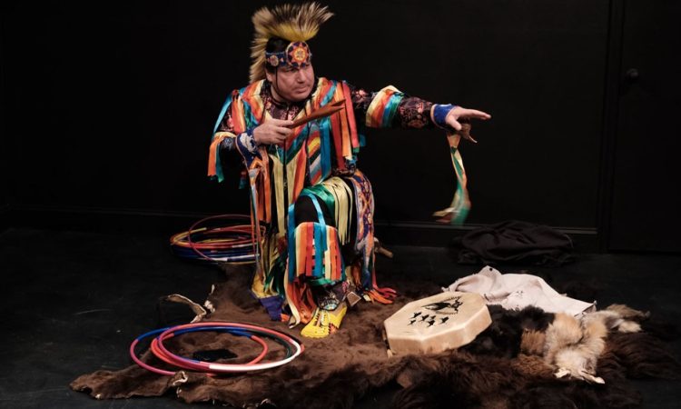 Delwin Elk Bear Fiddler kneels with one arm outstretched to the left and the other bent across his body holding a wooden pipe. Fiddler wears bright attire with multicolored strips of ribbons and a yellow feathered headdress. He kneels on a brown animal hide near a drum and several piles of plastic hoops in a blackbox theater.