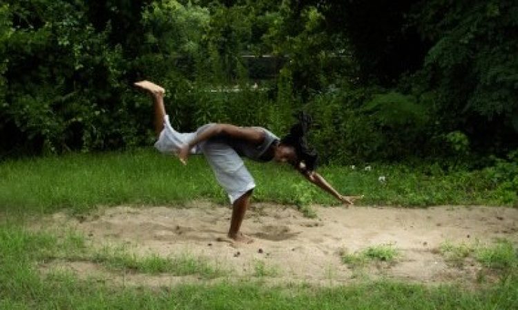 Marguerite Hemmings stands on one leg in active posture with one leg stretched behind them, arms outstretched in opposite directions, and torso parallel to the ground. They are surrounded by green trees and grass, standing on a patch of sand.
