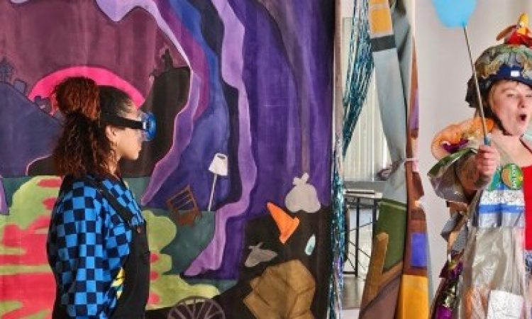 Two people stand in front of a curtain backdrop that’s painted like a trashed cave and swamp. One person is wearing a blue checkered longsleeve shirt with black overalls and big blue swimming goggles, holding the lid to a trash can. Another person is holding a blue fly-swatter in one hand and a toy stuffy in the other. They wear a costume made of trash and repurposed fabrics, and are standing in front of a large plastic trash bag.