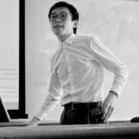 Mang Su (he/him) has short black hair and wears glasses. He is wearing a white button-down shirt, and is leaning on a table with a hand on his hip. The photo is in black and white.
