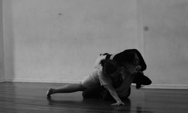 In a black and white photo, two dancers lay one on the ground, one on top of the other. The bottom dancer is supported by their outstretched hand on the ground, while the other touches the dancer's palm on top of them.