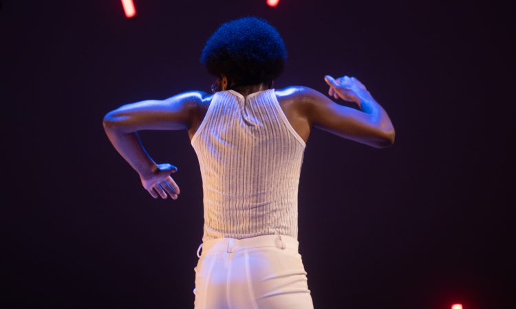 One figure with textured hair and deep brown skin lifts their elbows to shoulder height, their back to the camera, wearing all white.