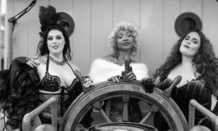 A black-and-white photograph shows three burlesque performers posing confidently at the wheel of Philly's historic ship, the Gazela Primeiro. Regal and composed, AshantiDoll stands directly behind the wheel in a sparkling gown with a white fur stole and short, curled blonde wig. On the left, Raven Nightingale wears a sequined top, fishnet stockings, and dramatic feathered sleeves, accessorized with a tall feathered headpiece. On the right, Miss Angie rocks a velvet corset-style gown with long gloves and voluminous curly hair as she tilts her head with a playful, bold look. The ornate ship’s wheel dominates the foreground,, anchoring the theatrical trio in a maritime setting that contrasts with their vintage burlesque styling.