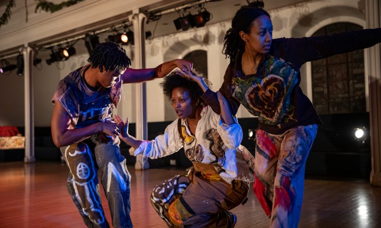 Performers Kris Lee and Stephanie Hewett lightly touch each others hands while they bend their knees, reach with their opposite hands, look away, as if about to leap or dash. In between them, performer AJ Wilmore is in a crouch, lightly touching the other two, looking into the distance. Their street clothes are colored with paint and what looks like white chalk, creating intricate and beautiful designs.