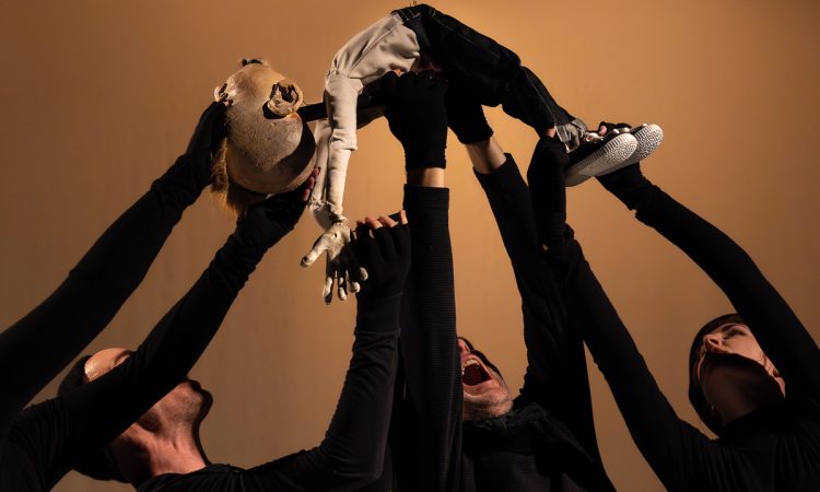 Four puppeteers dressed in black, standing close together with their arms raised as they support a small, human-shaped puppet above them. The puppet is shirtless and wears denim jeans and sneakers. It is held horizontally in midair, with its arms extended outward and its chest and abdomen lifted highest, creating a sense of suspension. The puppeteers are only partially visible, their faces and arms angled upward toward the puppet. A plain, warm-toned background and controlled lighting create strong contrast between the figures and the setting.