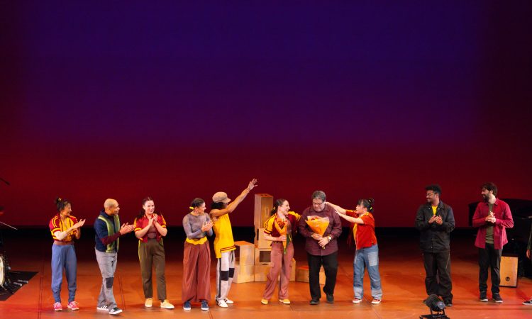 The image shows a group of performers standing in a line onstage, facing the audience. They are positioned in front of a red and blue gradient backdrop. Several people are clapping, as Ephrat Asherie and Arturo O’Farrill each hold a bouquet of flowers center-stage. Wooden boxes are stacked behind the group. Most performers wear casual, colorful clothing, and stage lights and instruments are visible at the edges of the stage.