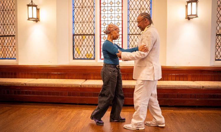 Two dancers stand facing one another, gently resting their hands on the other’s forearms. As one dancer steps forward, the other steps backward. The leading dancer is a woman, who wears a brightly colored headscarf, dangling earrings, a vibrant blue top, and stylishly baggy black jeans. The second dancer is a man, wearing an off-white linen suit, with his hair braided into neck-length cornrows. Both dancers are glancing towards the other’s feet as they move in sync through a partnered Tango sequence.