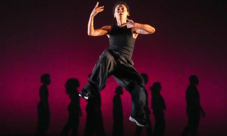 One dancer looks straight out into the distance, their face pinched in a look of concern, hands reaching back towards their chest. They are jumping up past a lineup of silhouettes imprinted on a blood red background.