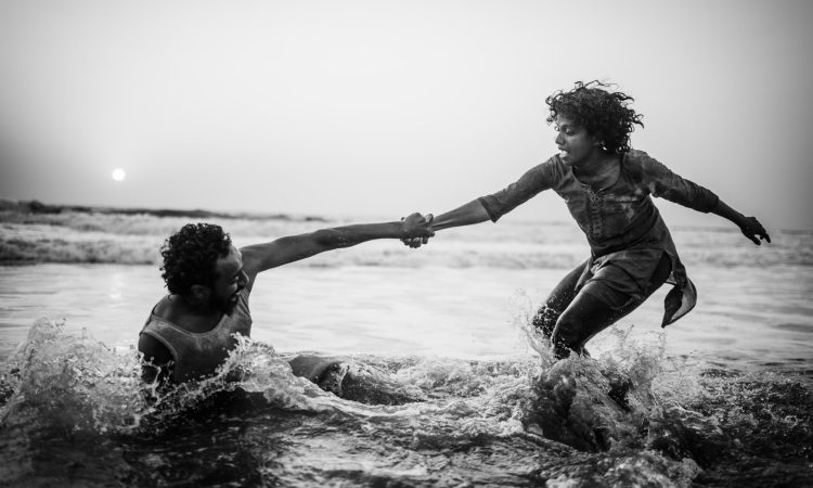 Two dancers practice the Contact Improvisation principle of weighing-out while in the shallows of the Arabian Sea at sunset. Small waves roll and crash in the background. The sun is a hazy dot in the upper left hand corner. Water crashes around the pair–a woman with short bouncing curls who stands, knees bent, and a man with short dark hair who presses one hand down into the water and sits on his side. Perhaps the wave has taken him out. The woman extends her left arm for balance as she reaches her right toward her partner. The two hold hands at the wrist as the water splashes around them. The photo is in black and white.