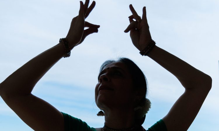 Nalini Prakash is backlit against a blue and white sky, where you can just see her head and arms. She holds two mudra shapes with arms bent, framing her head as she gazes towards her left hand. There is an airy lightness with her crisp gestures that frame a focused gaze.