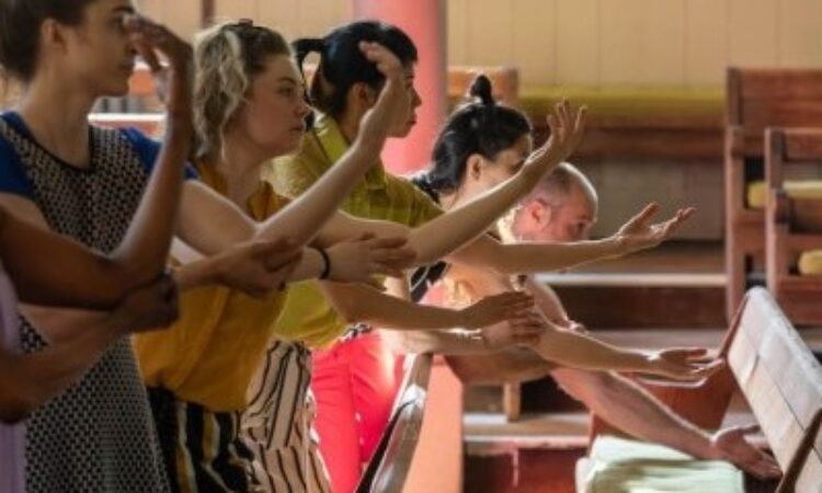The image shows a diverse group of people standing in a row inside a wooden bench, engaged in a canonic arm drop movement. They extend their arms forward with graceful hand gestures, focused on the action, creating a sense of unity and concentration.