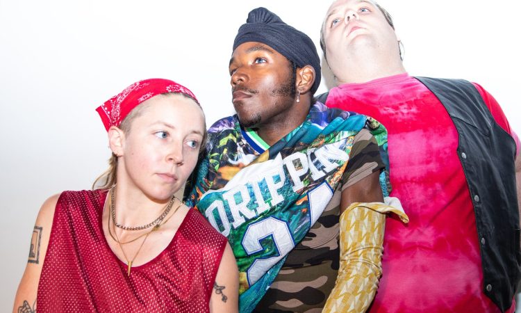 Three dancers in stand in a narrow diagonal against a stark white background. They all look off in different directions. On the left Annie Peterson wears a red bandana and a red pinnie gazes on a slight downward angle. In the center a Vitche-Boul Ra dons a colorfully patterned shirt, sets his sights up and to his right. On the right Zeze Schorsch in a red tie dye tee with a black vest overlain looks straight up, chin forward toward the camera.
