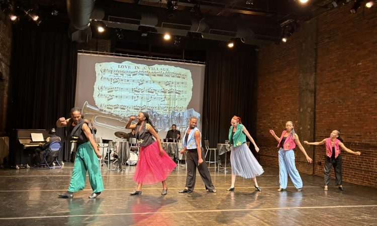 A group of six performers, four women and two men, are in a horizontal line dancing across the stage. They are dressed in colorful outfits that include pink skirts and colonial military styled vests in blue, pink, green, and black. Projected on the way is a background featuring a projection of sheet music titled "Love in a Village." A live five piece jazz band, including a piano, drums, a bass trumpet, and trombone, is visible behind the dancers upstage. The venue itself has a brick wall and dramatic lighting that aid in creating an engaging atmosphere.