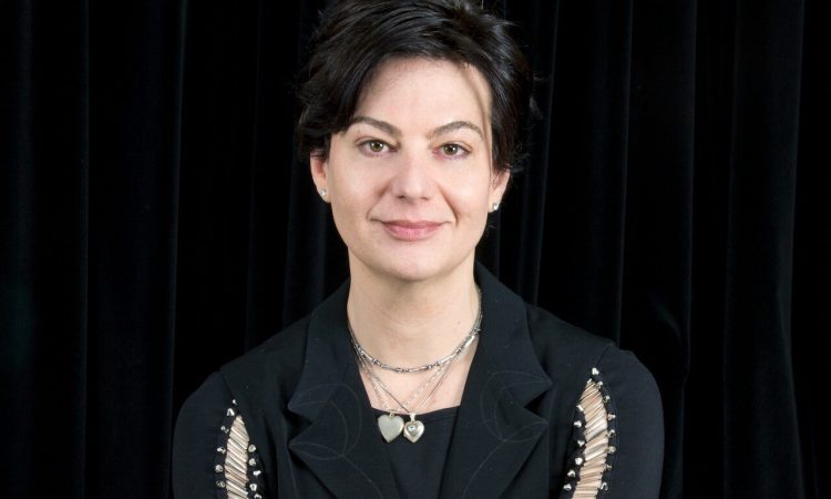 Richard Move sits facing the camera, a warm smile on their face. Their short wavy black hair matches the color of their long-sleeved top, which has thin lapels and light-colored stitches running down each arm. Two necklaces with heart-shaped pendants are visible.