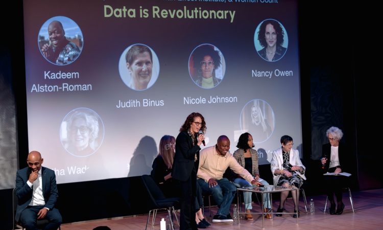 At the RISE 2025 Summit held at the New York Library for the Performing Arts, panelists for a discussion entitled “Data is Revolutionary” gather in black office chairs with silver legs in front of a large projector screen that reads “Dance Data Project, Harriet Tubman Effect Institute, & Women Count” in a pastel blue above the title “Data is Revolutionary” in a light yellow. Six circles containing the headshots of the panelists dot the rest of the projection. Amidst the other seated speakers, Nancy Owen stands holding a microphone in her right hand. She leans forward toward the audience, eyes alight, mouth open in speech that slips through an excited smile. In the lower left corner of the frame, a bald man in a crisp blue suit brings the crook of his thumb and second finger to his chin at stage right. He looks out toward the audience with his chair angled away from the six panelists.