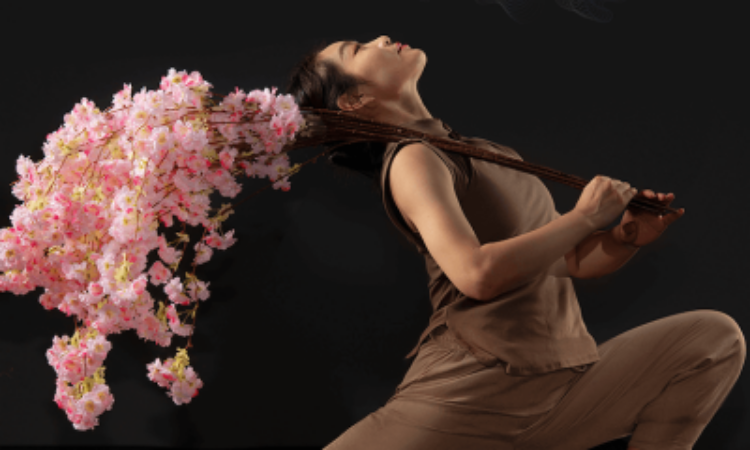 Weiwei Ma, dressed in a linen outfit, strikes a deep lunge against a dark background. With a branch of cherry blossom resting on her right shoulder, she arches her chest backward, gazing up toward the sky.