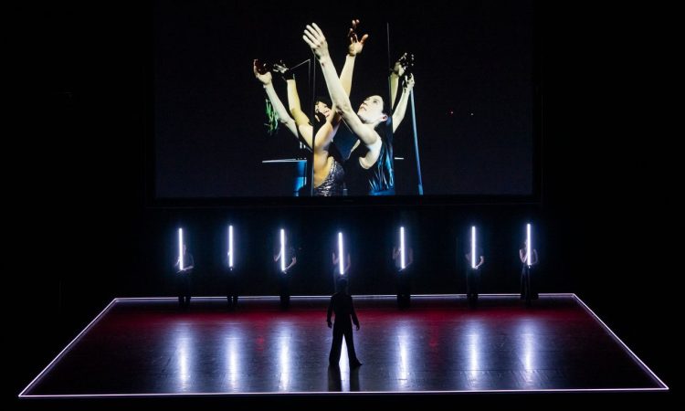 On a screen, Rachel Hutsell and Daphne Fernberger stand close together and reach up, crossing arms. The pair stands between a set of mirrors so their image reflects, causing the illusion of three sets of arms instead of one. On the stage below the screen, seven L.A. Dance Project ensemble dancers stand at attention, each holding a long, thin tube light vertically in front of them. At the front of the stage, a dancer in silhouette stands in a strong stance, feet wide, watching the scene unfold.