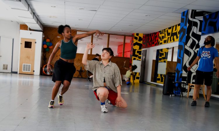 A spacious dance studio with a gray floor, mirrored walls, and colorful geometric murals is shown during a rehearsal for First Floor Spectrum. In the foreground, two people interact through expressive movement: one stands with an arm extended overhead while the other kneels and reaches upward toward the raised hand. Additional people are visible in the background practicing choreography, while another person stands near the right side of the room, directing. The studio contains chairs, exercise equipment, and a cluster of colorful balloons near the back wall. Natural light enters from windows along the left side of the space.