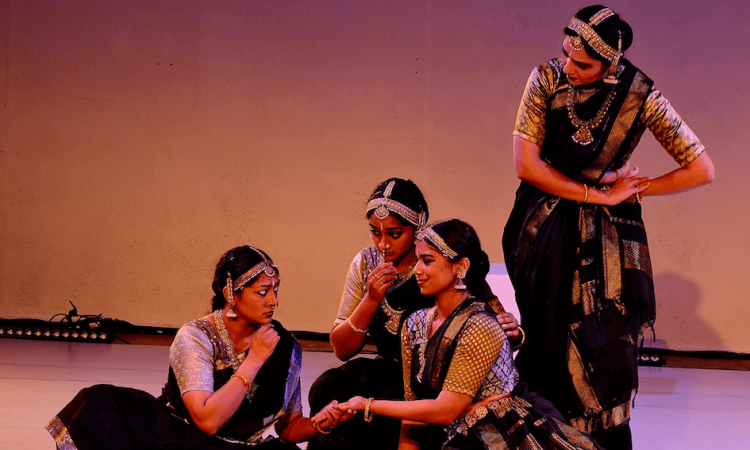 Four women gather onstage, three seated and one hovering above, creating a palm-reading tableau together. They wear black-and-gold metallic sarees and ornamental jewelry that adorns the crown of their heads. The dancer whose palm is being read wears a smug smile on her face while her two friends and the palm-reader seem puzzled, hands to their chins in contemplating her fate.