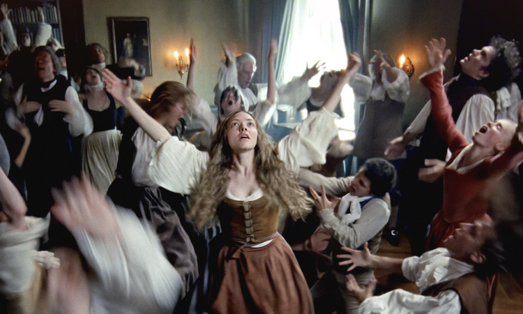 A scene from the 2025 film, The Testament of Ann Lee: Deep in the throes of ecstatic worship, Ann Lee (Amanda Seyfried), leads her congregation in an endurance challenge of dance, song and prayer. Men in powdered wigs and billowing sleeves and women in long dresses toss their heads back, open their mouths, and raise their arms. Two men near Lee kneel and stretch their arms out toward her. Lee is the only character fully in focus. Her blonde hair flies in front of her shoulders, chest heaving, as she raises her arms and eyes toward the Divine.