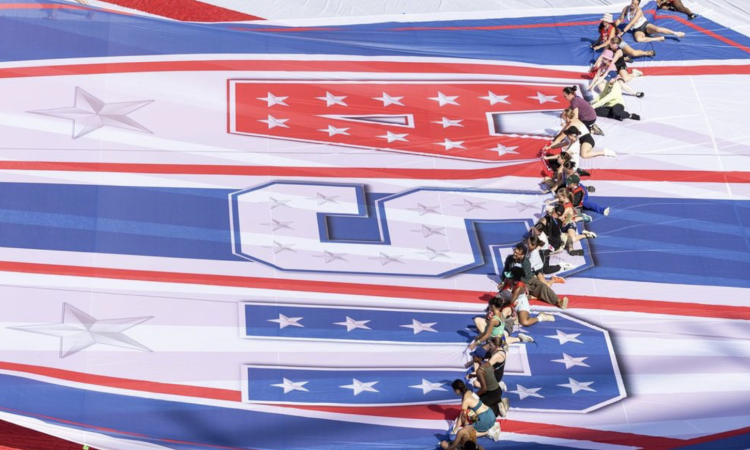 An overhead, high-angle view of a long line of people working together to unroll or pull a massive, vibrant patriotic tarp across a white field. The large-scale fabric features the letters "USA" written sideways in bold block lettering decorated with stars and stripes. The letter 'U' is blue, the 'S' is white, and the 'A' is red. Photo credit: Balich Wonder Studio.