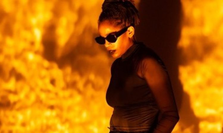 Angie Pittman stands with her hands in her pockets looking downward. She wears black sunglasses with her locs tied into a high ponytail. Pittman is doused in a textured golden amber light.