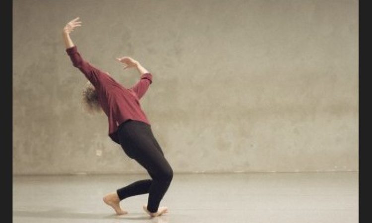 Positioned slightly to the left of center of the image, Lisa Kraus falls to the side over one ankle. Leaning back onto the ball of her other foot with her arms raised upward, her head lifts back, and her hair flies behind her. She is in a gray space wearing black leggings and a maroon long sleeve shirt. The photo is from 2025 and was inspired by the black and white photo by Lois Greenfield, described below under the heading “Homepage Image Description.”