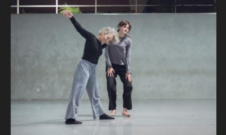 Smiling with her left foot stepped forward and right arm lifted, Lisa Kraus angles her weight lightly toward Laure de Dietrich. Kaus’s left hand rests on de Dietrich’s right arm. Both dancers have their heads inclined slightly to the left. de Dietrich may be descending from a small jump – both of her hands rest on her thighs, fingers spread, palms face-down, knees with a micro bend and feet supinated just off the ground. The duet takes place at Tictac Art Centre.