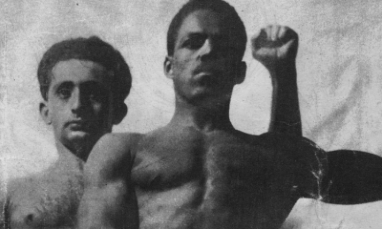 Hy Boris and Add Bates, two lean and muscular male dancers, stand shirtless and flex their biceps in a pose reminiscent of Edith Segal’s Black and White. Their faces are powerful and commanding.