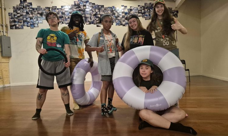 Image Description: Five young would-be campers, mostly smiling, with goofy patterned T-shirts standing in socks on a wood floor displaying supportive thumbs up. One dancer is seated, their face framed by a round lavender and white floaty.