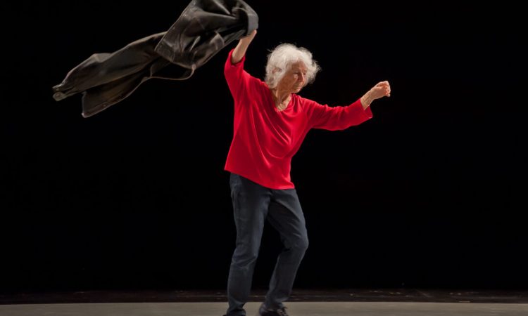Standing, Simone Forti lifts both of her arms. She wears a foream-length red shirt, and she has short white hair.