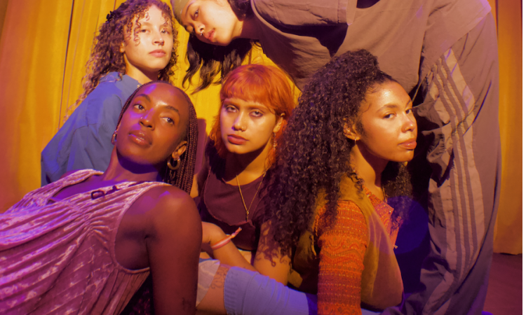 With serious expressions and close proximity to one another, five dancers sit, lean, stoop, and rest casually.