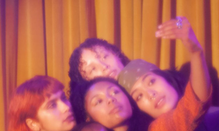 Five dancers lean toward one another, and one holds up an invisible phone as though they’re pantomiming taking a selfie. They all look serious. The image is soft and grainy.