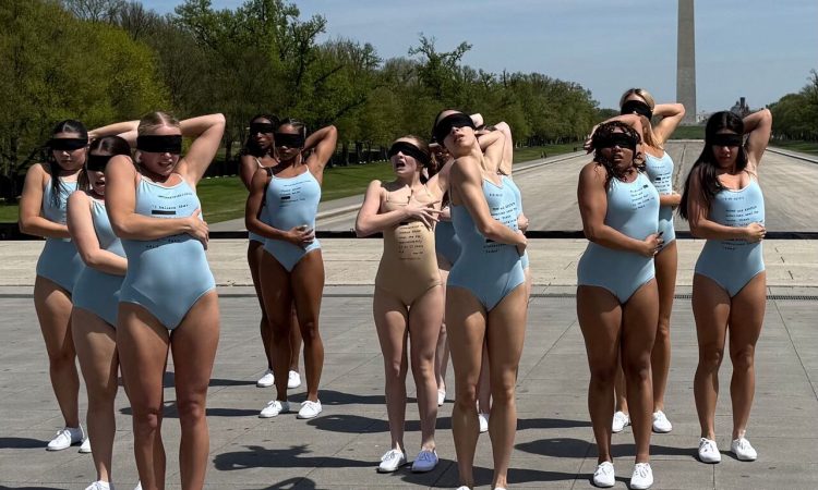 An ensemble of dancers stand upright wearing form-fitting leotards and lace-up white shoes. All have a sheer black cloth covering their eyes. Their left arm reaches behind their heads, elbows flexed, and their right arm snakes across various places of their waist, ribcage, and back. All of the dancers look in varying states of unease.