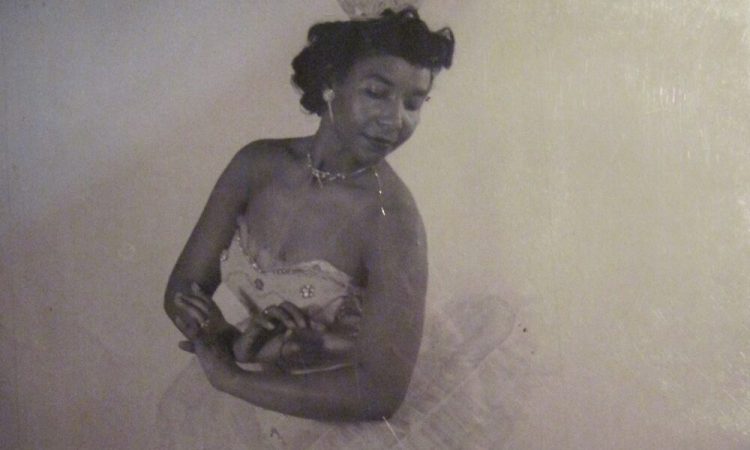 Black and white photo of Sydney King posing in a tutu and matching crown. Her arms are gently crossed in front of her torso and her head tilts back as if looking over her shoulder. King wears point shoes and stands in tendu derriere, one leg extended behind her.