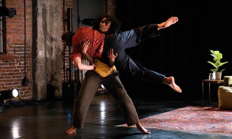 Chloe Marie and Tamar Gutherz are captured on stage mid-stunt in this action shot. Chloe is standing upright grasping onto Tamar who is horizontal in their arms with legs extended long. There is an area rug and an end-table with a potted plant in the background.