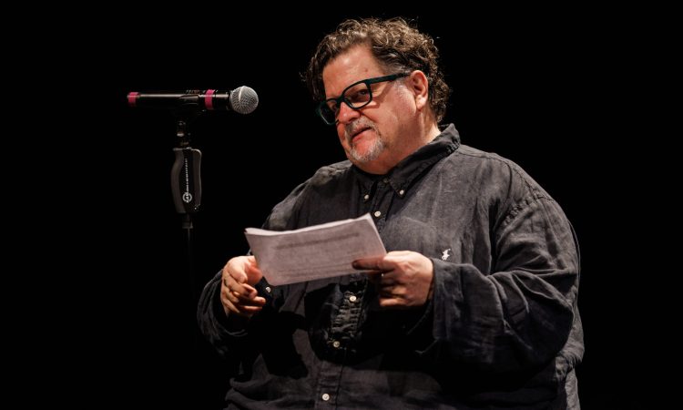 A man (Tere O´Connor) wearing a dark button-down shirt sits in a chair, a microphone in a stand to his right. His head is tilted slightly to one side, and he wears black-rimmed glasses and has a goatee speckled with gray. In his hands he holds some papers, as if he is reading something of importance.