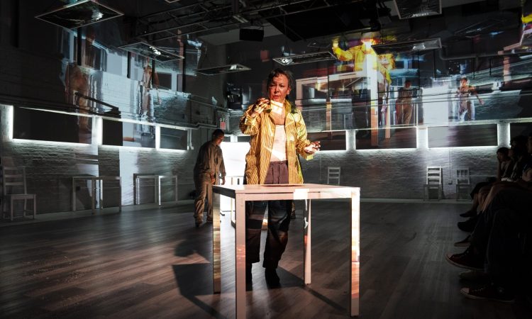 maura nguyễn donohue stands behind a white table, with overlapping projections of herself and the room around her covering her face and body. She wears a mustard yellow button-down shirt and holds a small object out in front of her, looking at it with curiosity. Shannon Yu is standing, slightly slumped, behind nguyễn donohue. Yu wears a monochrome grey outfit, and gently shrugs her head and shoulders forward, curving her upper spine.