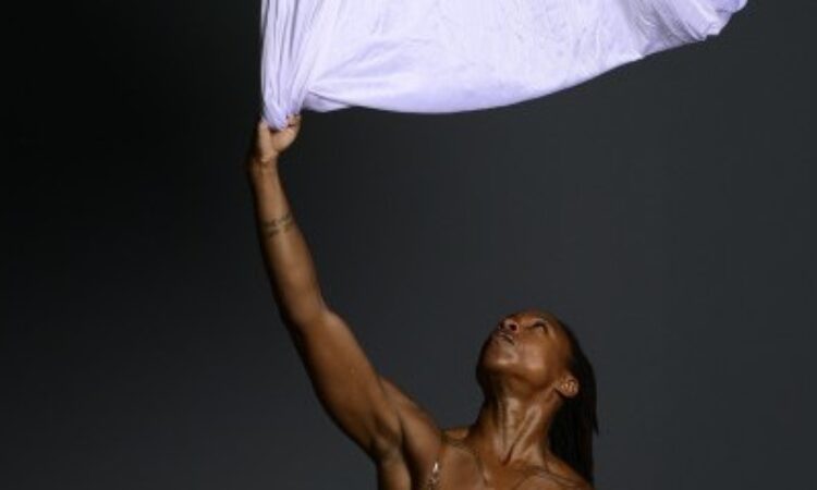 Joi Cox, a muscular female dancer, reaches and looks toward the ceiling from which thick white cotton fabric hangs.