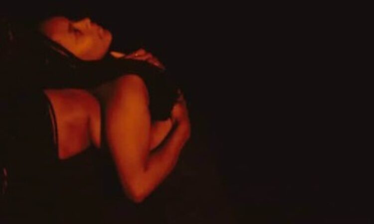 A dancer with braids leans against the shoulder of another dancer in a dimly lit room with a red hue. Both wear black and seem to be enjoying each other's company.