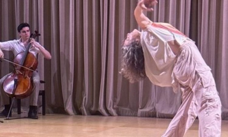 Tristan Price plays the cello seated on stage with a black microphone positioned in front of him. He wears black shoes, khaki pants and a white button-down shirt. He looks directly at dancer and his mother, Megan Bridge, who is dancing next to him with arms outstretched in his direction. Bridge wears a white top and grey cargo pants with sneakers.