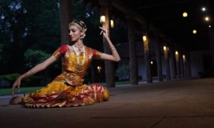 A woman sits on the stone floor of a long veranda at dusk, illuminated by hanging lanterns. The warm light makes her golden costume glow. Resting on her left hip, her right leg extends behind her, half bent. She looks down her extended right arm, letting her fingers drop toward the floor, while her left hand, at head level, pinches some fingers into her palm, leaving her ring and pinky fingers reaching up.