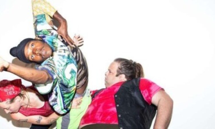 Three dancers in dynamic positions pose against a stark white background. On the left Annie Peterson wears a red bandana and a red pinnie is hunched over looking down. In the center a Vitche-Boul Ra dons a colorfully patterned shirt, has one arm akimbo and the other reaches out towards the lens. On the right Zeze Schorsch in a red tie dye tee with a black vest overlain leans back and looks towards the other dancers.