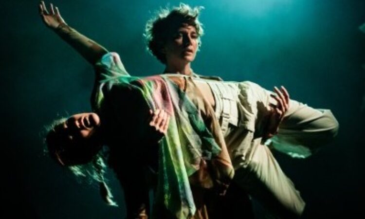 One woman, Zoe Rabinowitz, with green hued lighting behind her, is carrying horizontally across the front of her torso, Mary McGrath, who is lit with green and red hues across her outstretched body. Rabinowitz gazes forward beyond McGrath, who lies motionless on her back across the supporting arms of Rabinowitz.