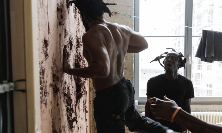 In a scene from Dambudzo, tyroneisaacstuart punches red clay into a cardboard wall, face turned away from the camera, as Tatenda Chabarwa looks on at this scene, face aghast in horror. Both are Black men outfitted in plain black clothing that both contrasts their dark brown, Black skin tone and captures remnants of red clay that spatters off the wall.
