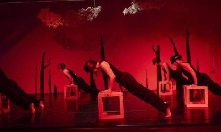 A group of dancers wearing all black, with old-fashioned wartime helmets assume a slanted plank position with arms leaning into wooden cubes. A red light encapsulates the space, as large black spikes climb upwards along the back wall and two permeable sculptures are hung from the ceiling.
